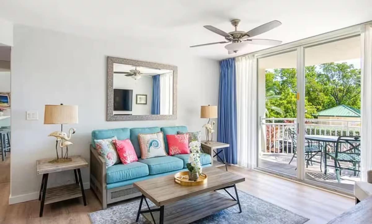 A bright living area features a plush blue sofa adorned with colorful throw pillows. A coffee table sits in front of the sofa, surrounded by light wood accents. Large glass doors lead to a private balcony, inviting natural light and views of greenery.