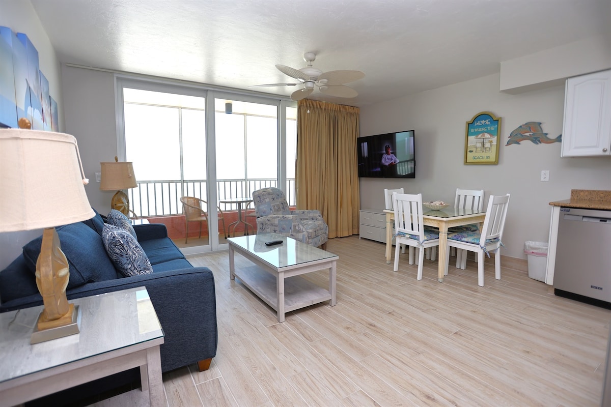 A spacious living area features a navy blue sofa with light accent pillows, a coffee table, and a flat-screen TV. A dining table with white chairs is positioned nearby, complemented by light wood flooring. Large sliding glass doors allow natural light and provide access to the balcony.