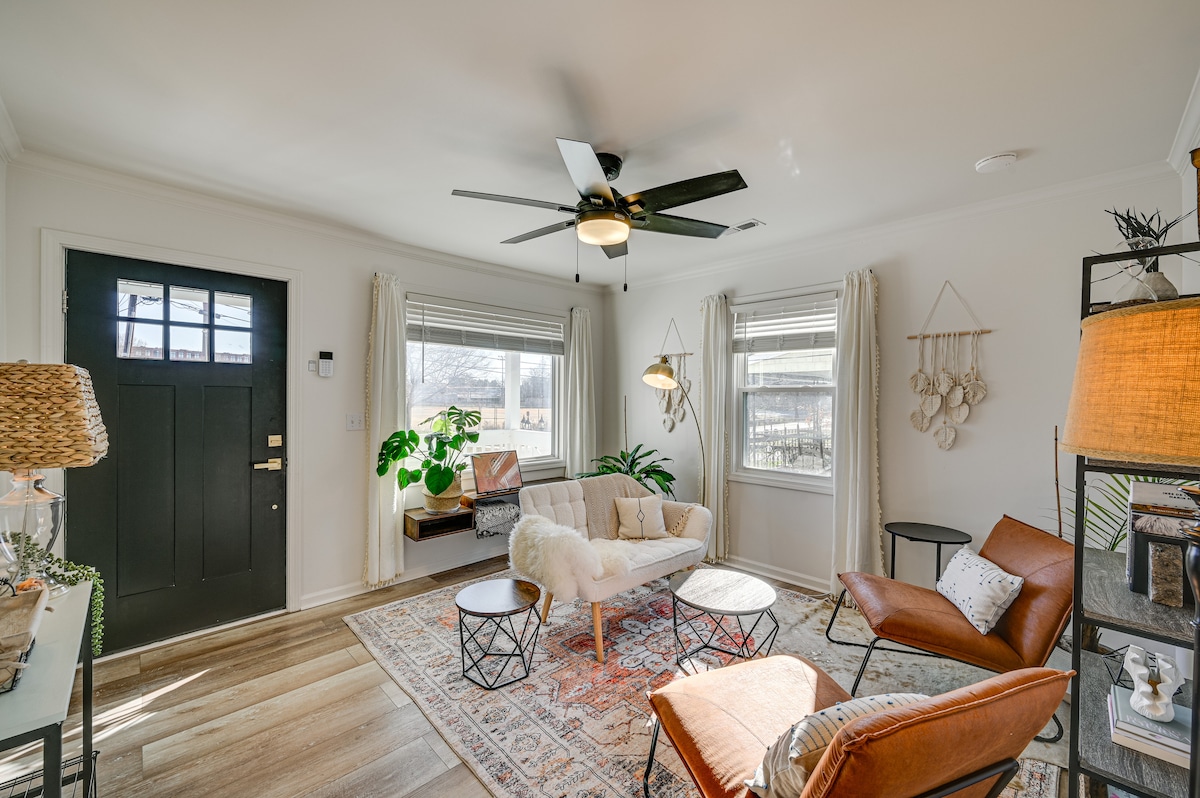 A bright living area features a comfortable seating arrangement with a spacious couch and two accent chairs. Natural light enters through large windows, highlighting a stylish ceiling fan and warm wood flooring. Decorative elements include an area rug and various plants integrated into the decor.