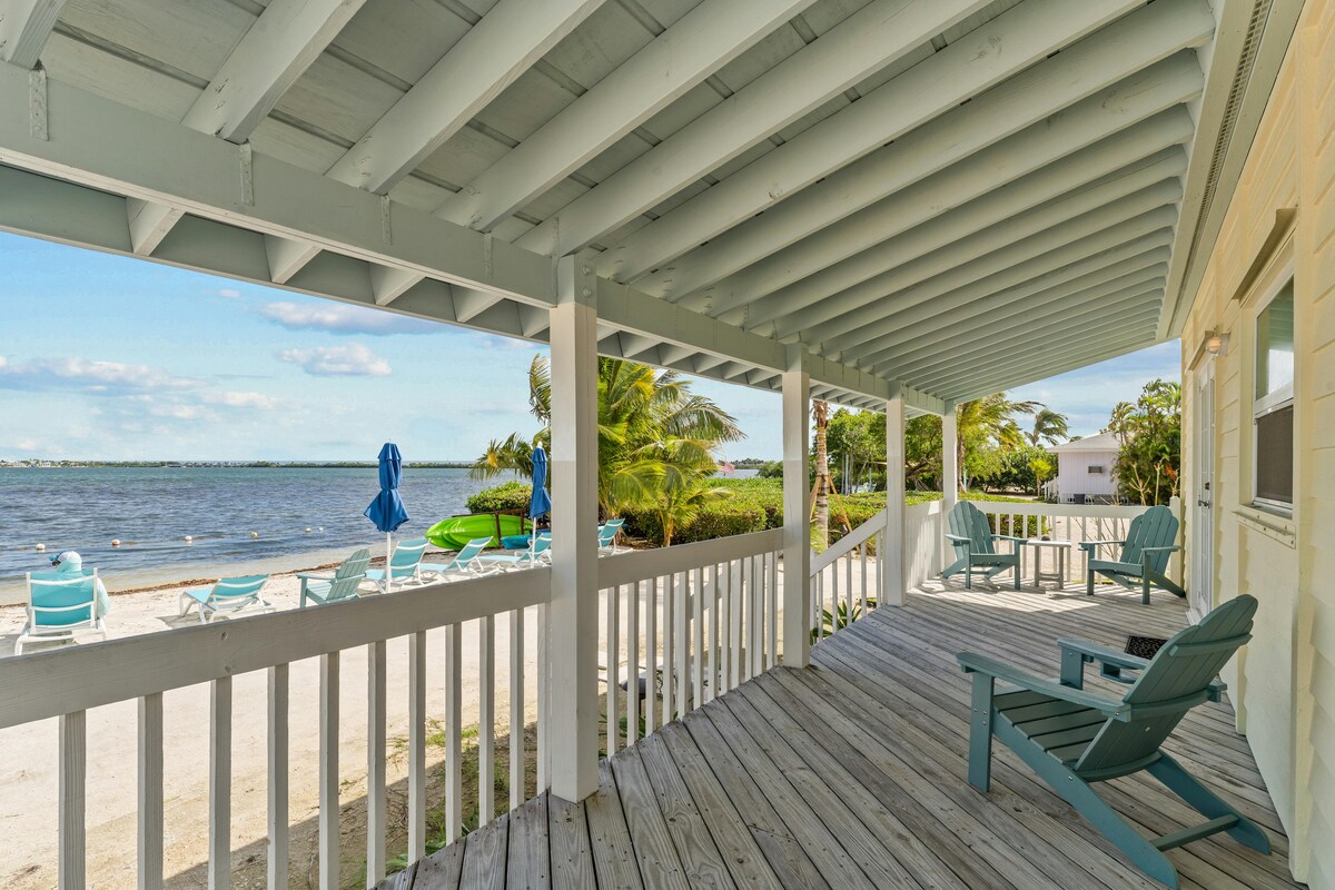 A covered porch overlooks a peaceful waterway, featuring multiple blue outdoor chairs and umbrellas. The wooden deck is illuminated by natural light, with gentle waves visible in the distance among palm trees, creating a serene outdoor space ideal for relaxation.