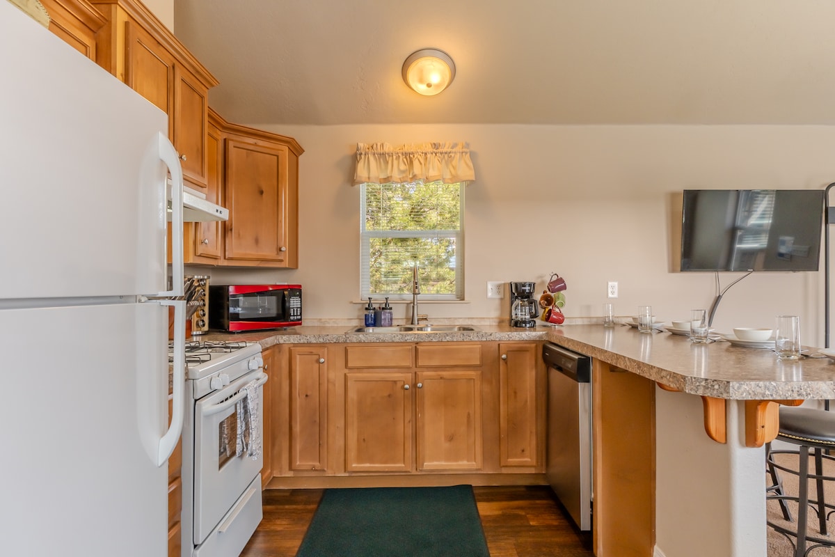The kitchen area is equipped with wooden cabinetry providing ample storage. A stainless-steel dishwasher and refrigerator complement a gas stove and microwave. A countertop with seating is visible, along with a large window allowing natural light to fill the space.