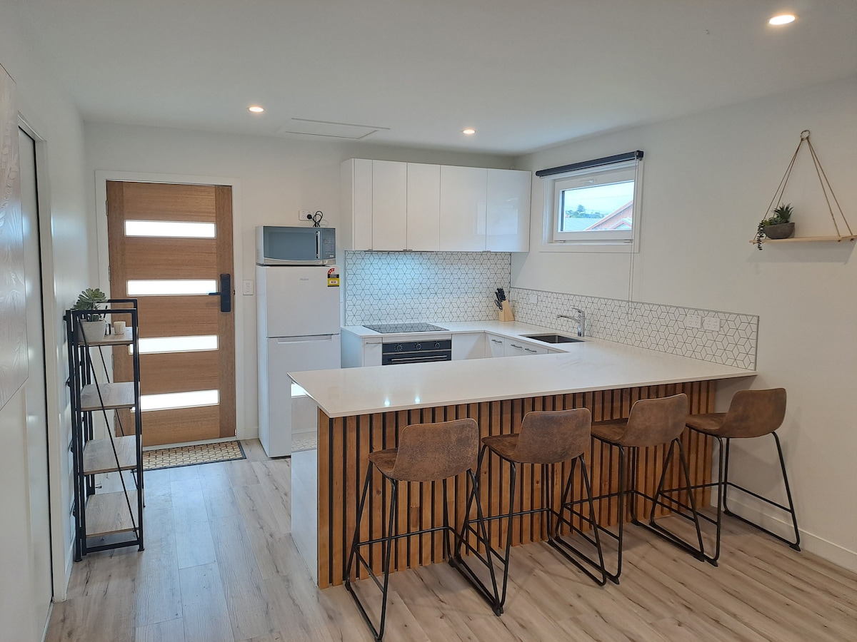 The kitchen area presents a modern design with white cabinetry and a large breakfast bar featuring four high stools. A refrigerator is visible, alongside a sink and stovetop. Natural light streams in from the window, illuminating the light wood flooring.