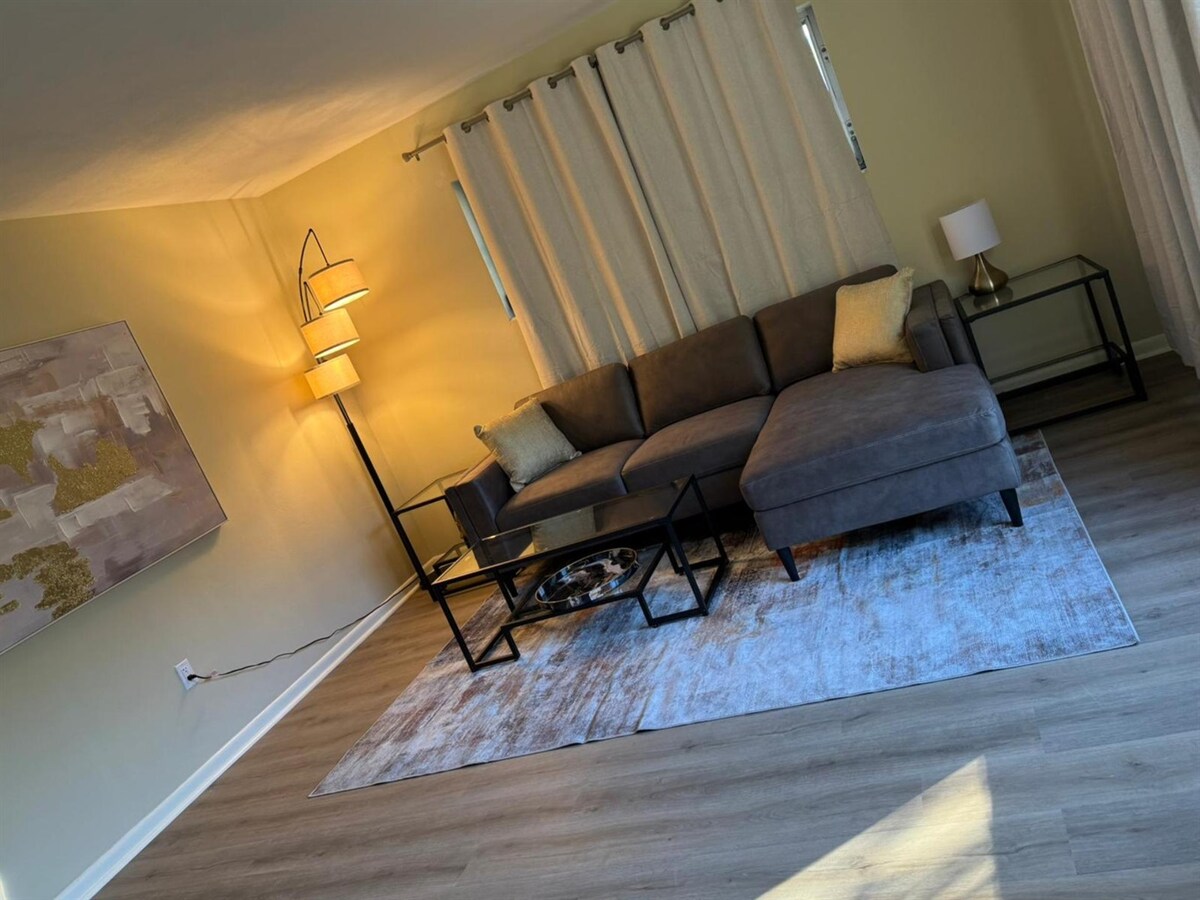 A comfortable living area is shown featuring a sectional sofa with soft throw pillows. A glass coffee table rests on a textured area rug. Natural light enters through large windows covered by light curtains, while a floor lamp adds warm illumination to the space.