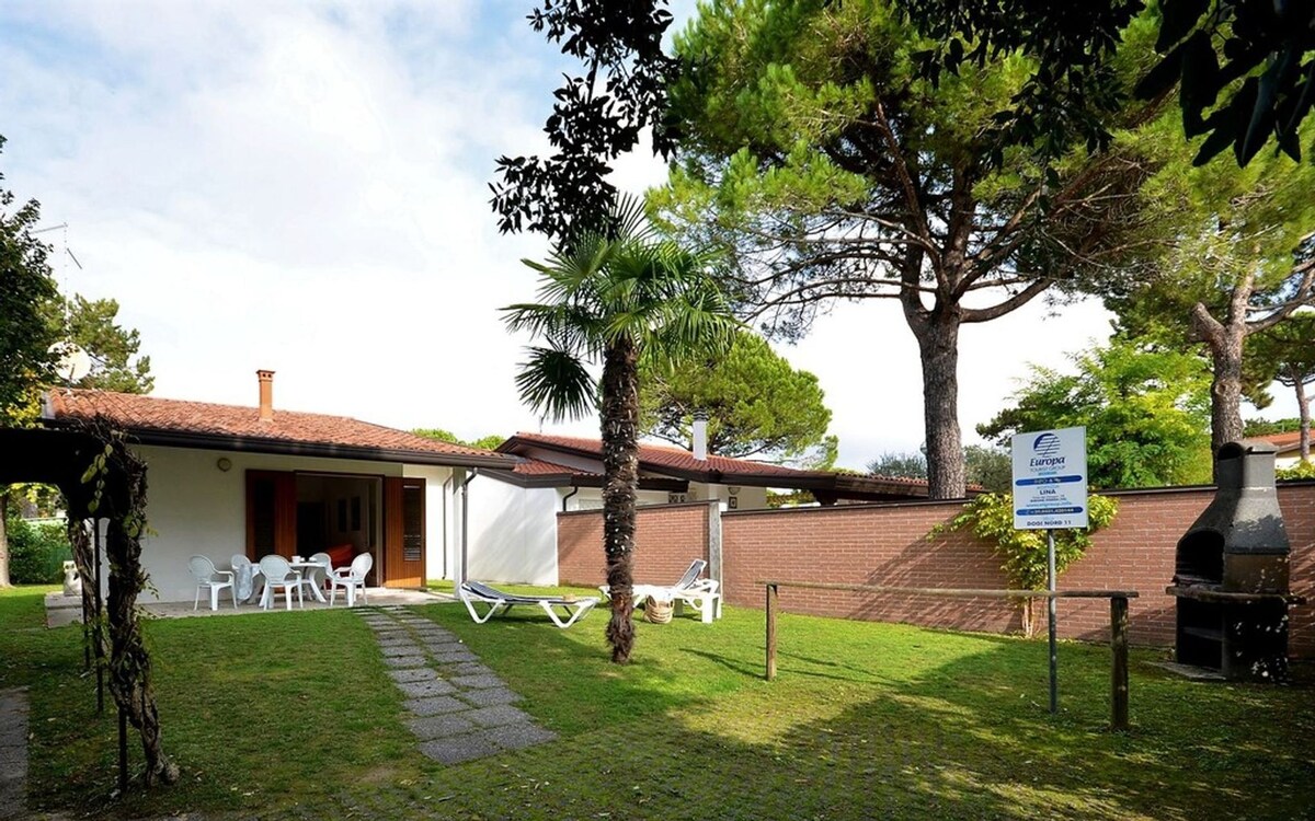 A spacious outdoor area features a well-maintained garden with green grass and trees. Several lounge chairs are positioned for relaxation. The villa’s entrance, marked by large windows, is visible along with a barbecue area, providing an inviting space for outdoor gatherings.