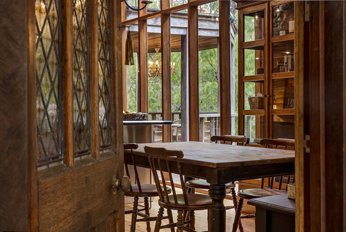 An inviting dining area features a rustic wooden table surrounded by several chairs. Large glass doors provide views of the surrounding trees and let in natural light. Shelving units displaying various items offer a cozy, personal touch to the space.
