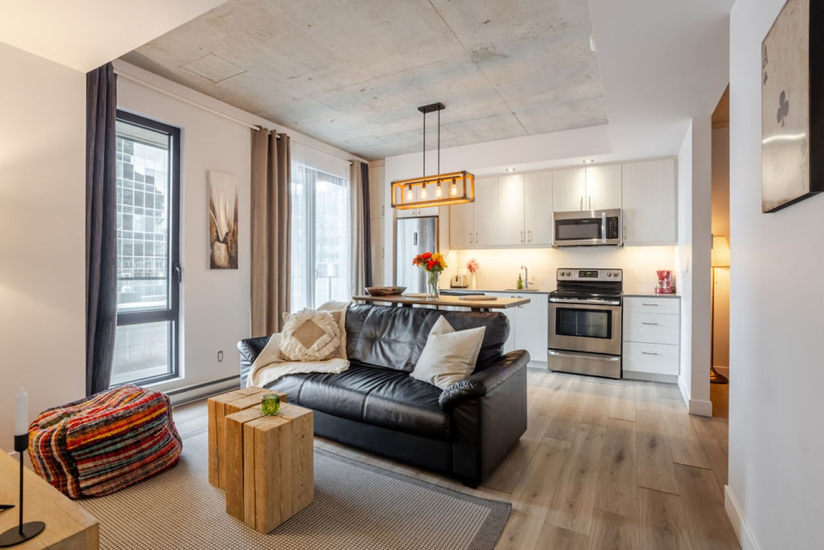 A welcoming living area features a leather sofa adorned with decorative pillows, alongside a small wooden coffee table. Bright natural light enters through large windows with light curtains, illuminating the modern decor. The open kitchen is visible, equipped with stainless steel appliances and a stylish light fixture above.