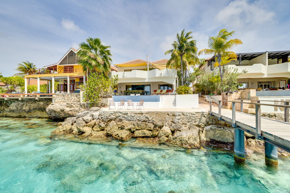 A welcoming villa is set on rocky coastal terrain, with lush palm trees surrounding the property. A private pier extends into clear blue waters, while open terraces invite relaxation. The structure features multi-level outdoor spaces, perfect for enjoying the serene seafront views.