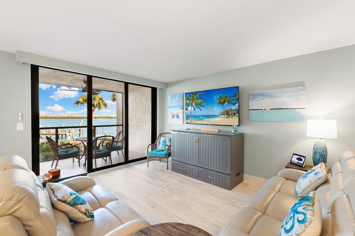 A spacious living area features two light-colored sofas adorned with decorative cushions. A large flat-screen television is mounted above a console, and light streams in through sliding glass doors that open to a private lanai overlooking the waterfront.