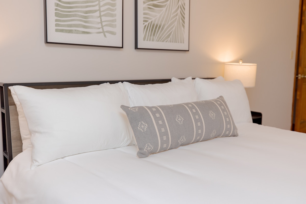 A spacious bed is presented with crisp white linens and decorative pillows. Framed botanical prints are hung above the headboard, while a soft lamp emits gentle lighting from the bedside, creating a calming ambiance.
