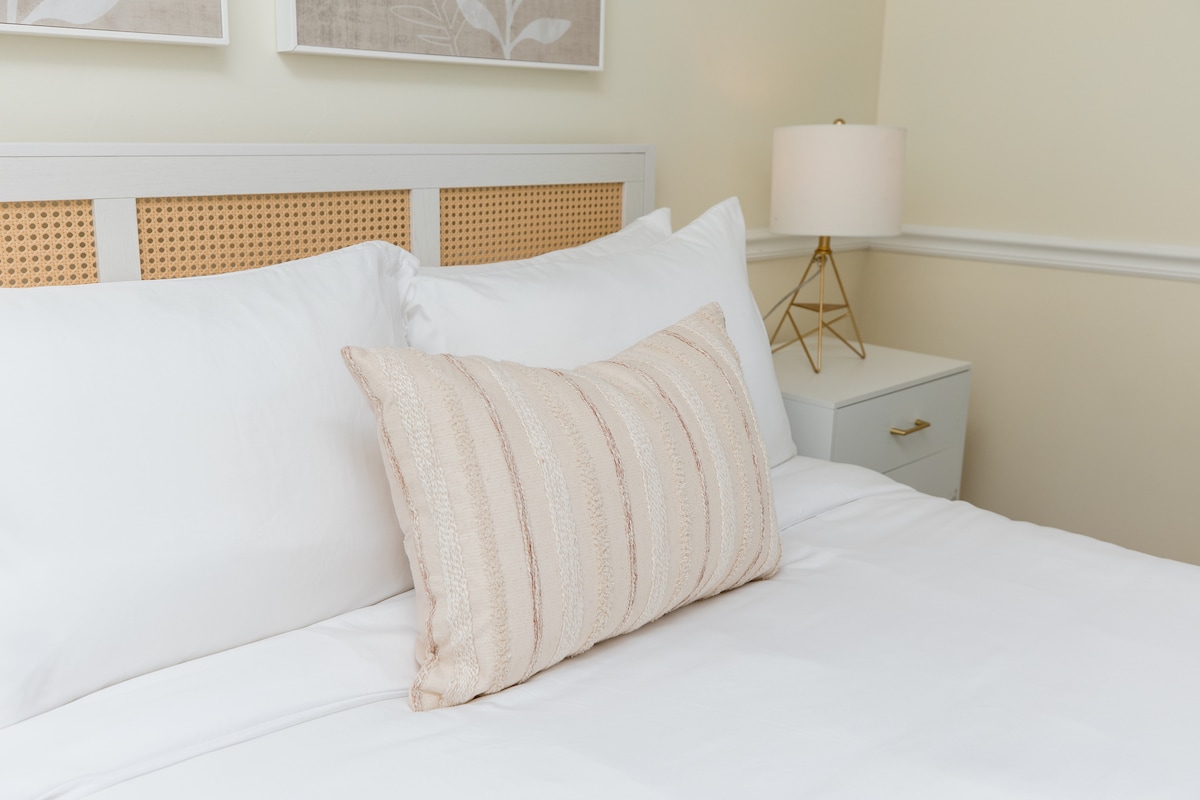 A well-made bed features crisp, white linens with a decorative striped pillow positioned at the center. Soft light is provided by a modern lamp set on a nightstand beside the bed, complementing the warm, neutral tones of the room.