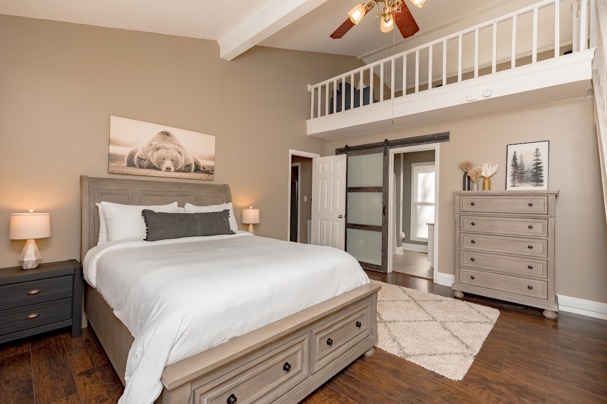 The spacious master bedroom features a queen bed with a comfortable, neutral-colored bedding set. Light wood furniture, including a dresser and bedside tables, complements the decor. A loft space is visible above, with bright natural light streaming through nearby windows.