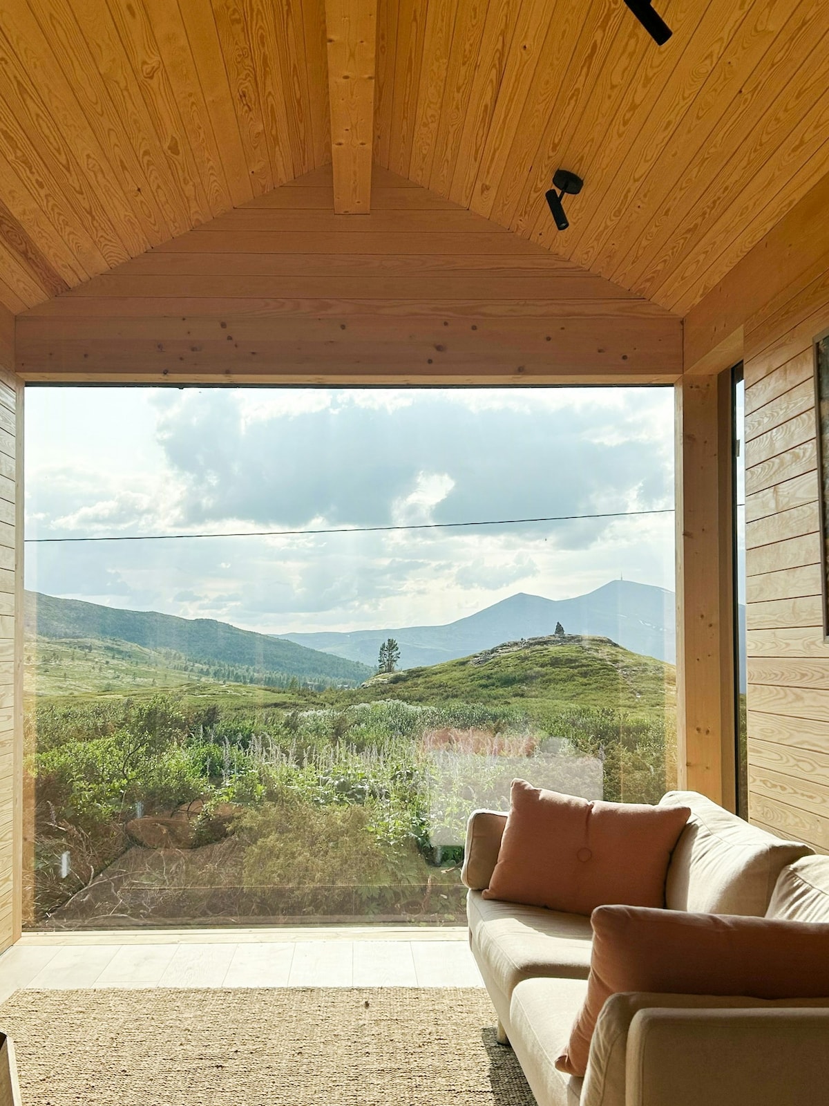 A cozy seating area features a light-colored sofa positioned against large glass windows, offering expansive views of rolling hills and distant mountains. The natural wood paneling of the walls adds warmth, complementing the serene landscape visible outside.