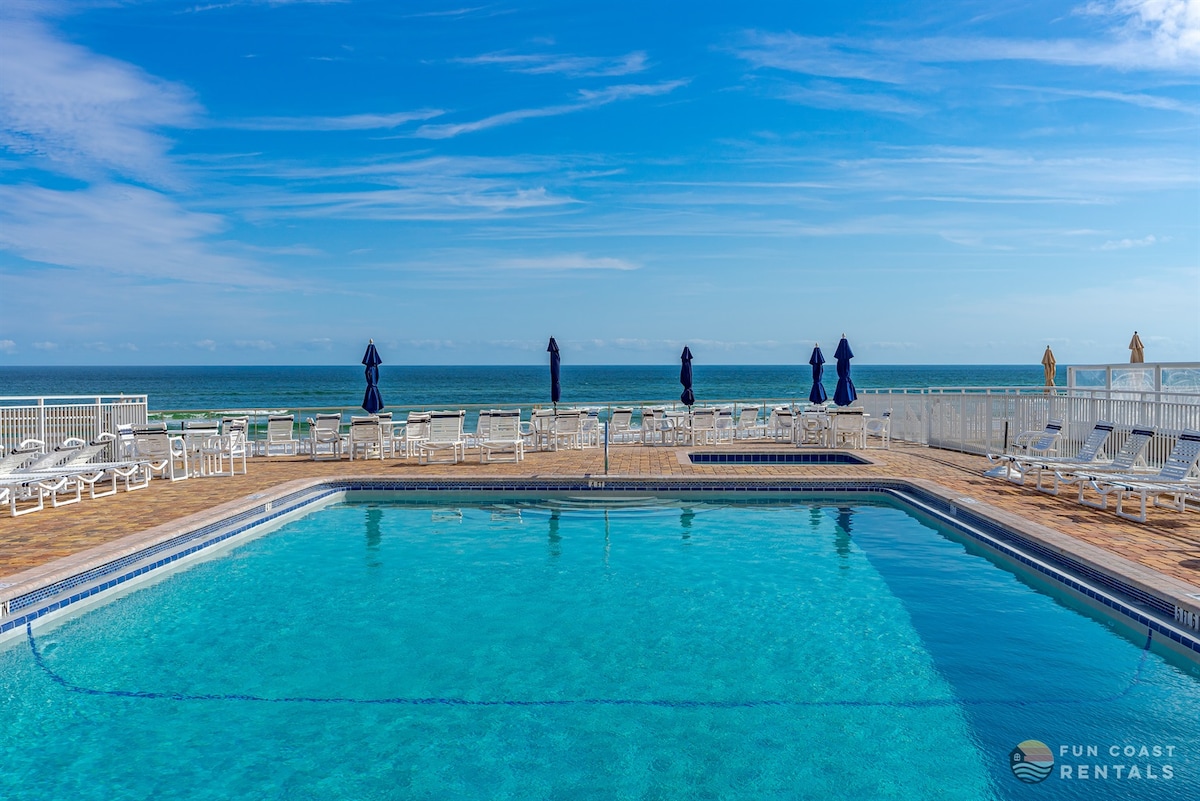A clear view of the sparkling pool surrounded by lounge chairs is presented, with umbrellas positioned for shade. The ocean is visible in the background under a bright blue sky, highlighting a serene and inviting atmosphere for relaxation.