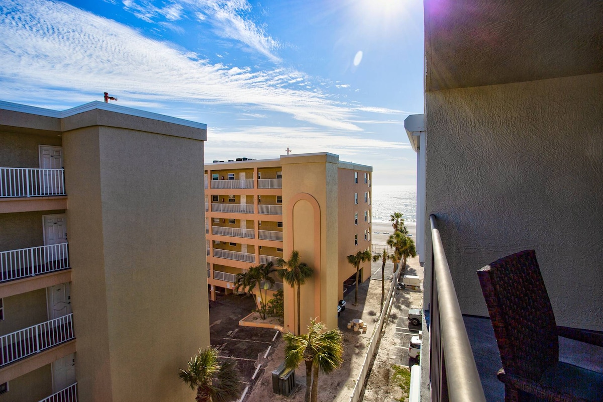A view from the top-floor balcony captures the surrounding beachfront buildings and the expanse of the sky. Sunlight reflects off the ocean in the distance, with palm trees framed below, creating a serene atmosphere.