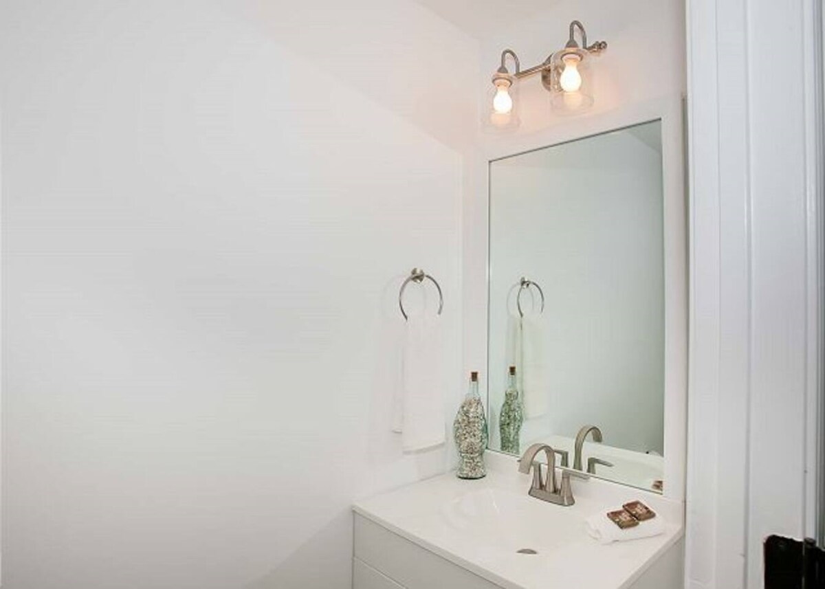 A bright bathroom features a minimalist design with a white vanity and sink. A large mirror reflects the room, complemented by two light fixtures. A hand towel hangs on a wall-mounted ring, and decorative glass bottles are positioned beside the sink.