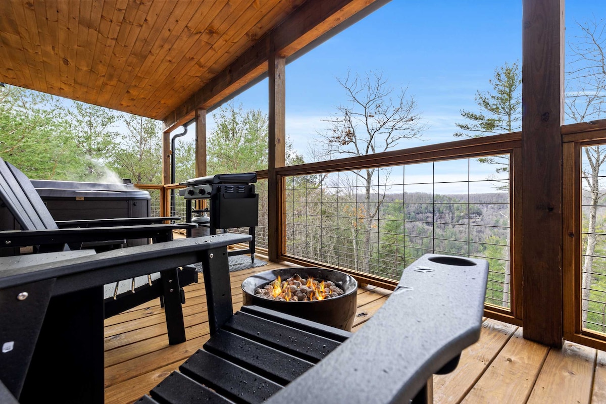 A covered back deck features two Adirondack chairs and a propane firepit, overlooking scenic views of the surrounding trees and hills. A hot tub is visible in the background, with steam rising gently, adding to the relaxing ambiance of the outdoor space.