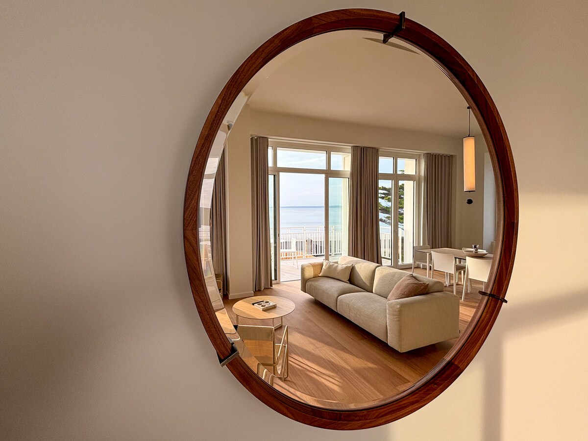 A circular wall mirror reflects a spacious living area that showcases a light-colored sofa, a small coffee table, and a dining set. Large windows provide a view of the sea, allowing natural light to fill the room, highlighting its welcoming ambiance.