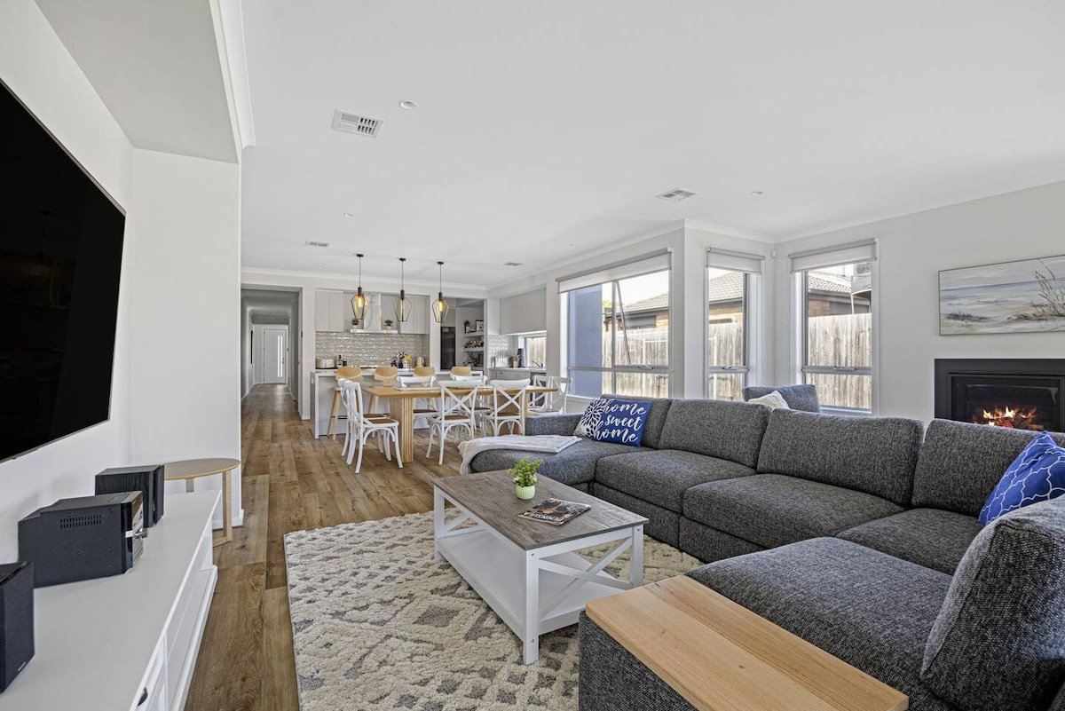 An open-plan living area features a spacious grey couch arranged around a coffee table. A large TV is mounted on the wall, with a cozy gas fireplace nearby. Natural light floods in through large windows, highlighting smooth wooden floors and a dining area in the background.