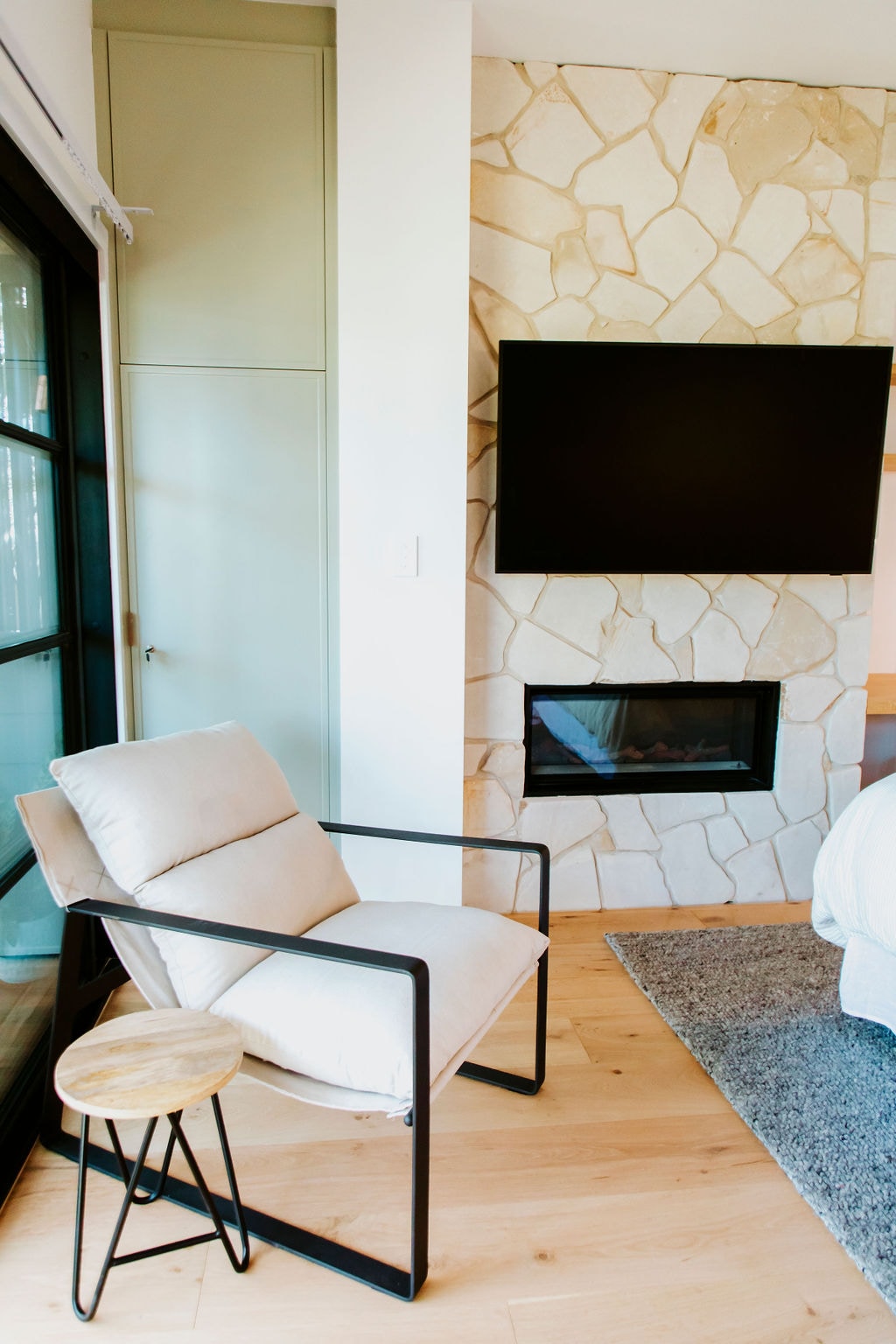 A cozy corner of the studio presents a sleek chair with a wooden side table beside it. The stone feature wall serves as a backdrop to the television mounted above a stylish fireplace, creating an inviting atmosphere.