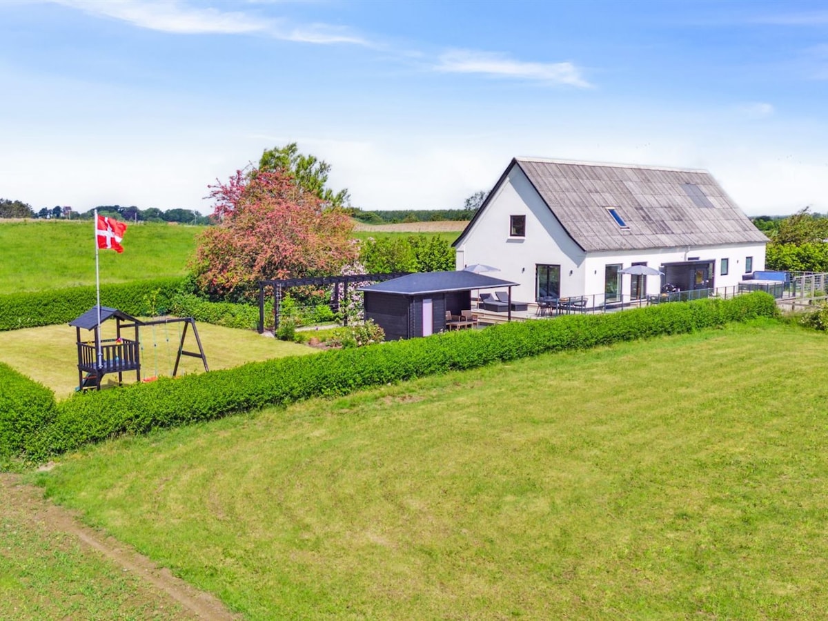 An expansive green lawn surrounds the property, with a children's playground featuring a swing set on one side. The well-kept garden is lined with hedges and a tree, while the white single-family house, with a sloped roof, is visible in the background.