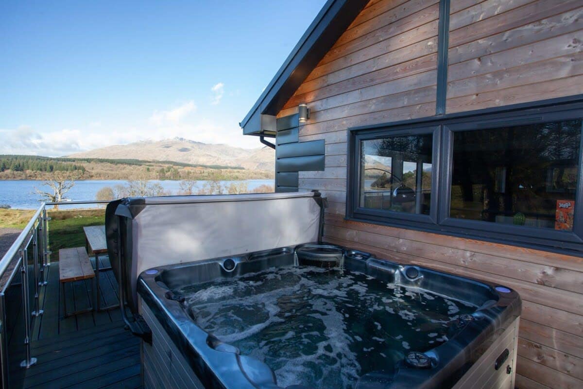 An outdoor hot tub is situated on the deck of the lodge, offering scenic views of Loch Awe and the surrounding landscape. The modern wooden paneling of the lodge complements the tranquil setting, with a spacious dining table positioned nearby for al fresco meals.