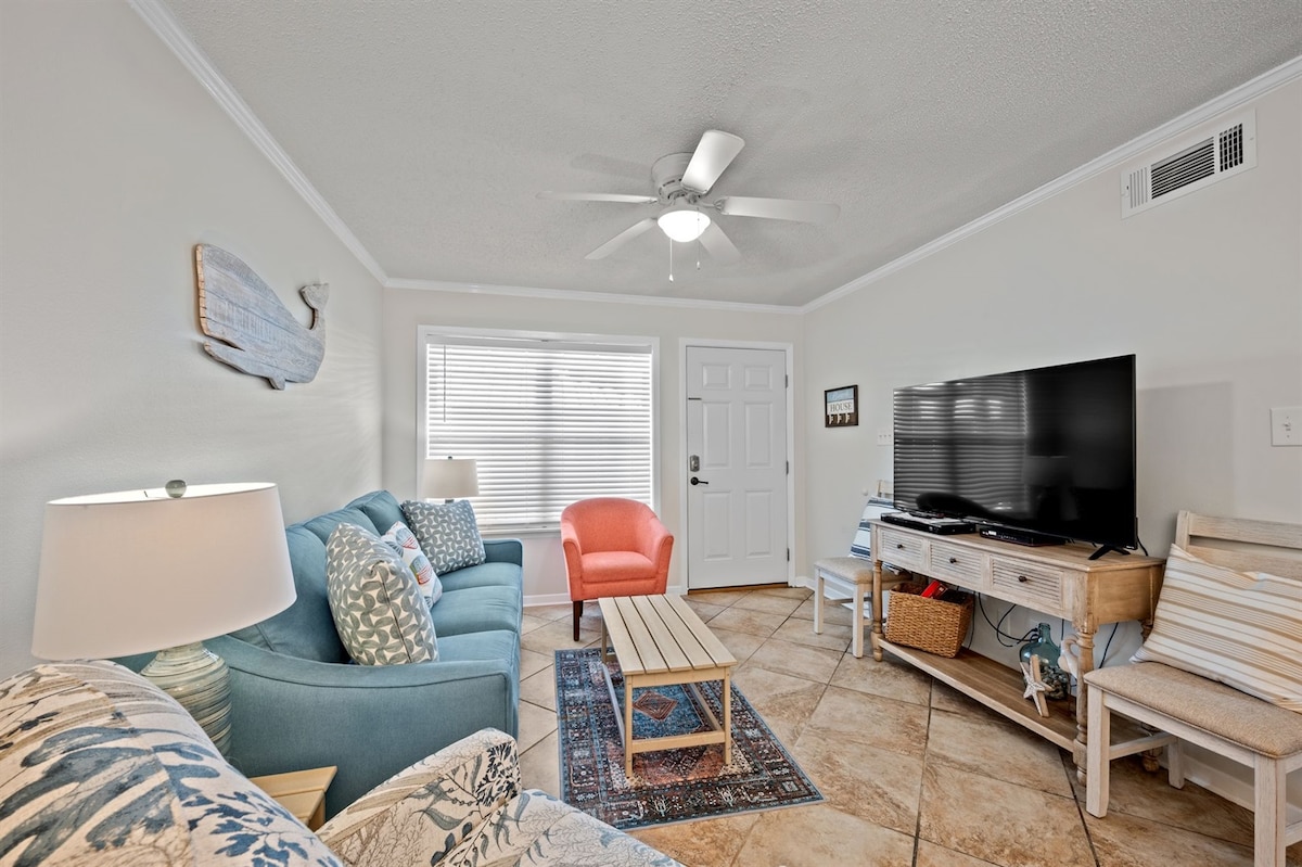 The living area features a cozy arrangement with a blue sofa and an armchair in coral. A light wood coffee table sits in the center, accompanied by a decorative rug. Natural light enters through the window, while a ceiling fan provides airflow. A flat-screen TV is mounted on the wall.