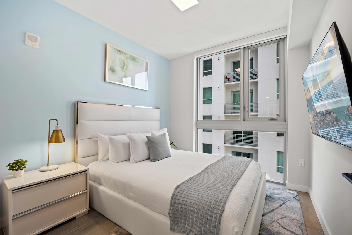 A serene bedroom features a cozy queen bed with plush linens and decorative pillows. A bedside lamp provides soft lighting. Large windows allow natural light and offer a glimpse of the outdoor view. A sleek television is mounted on the opposite wall for entertainment.