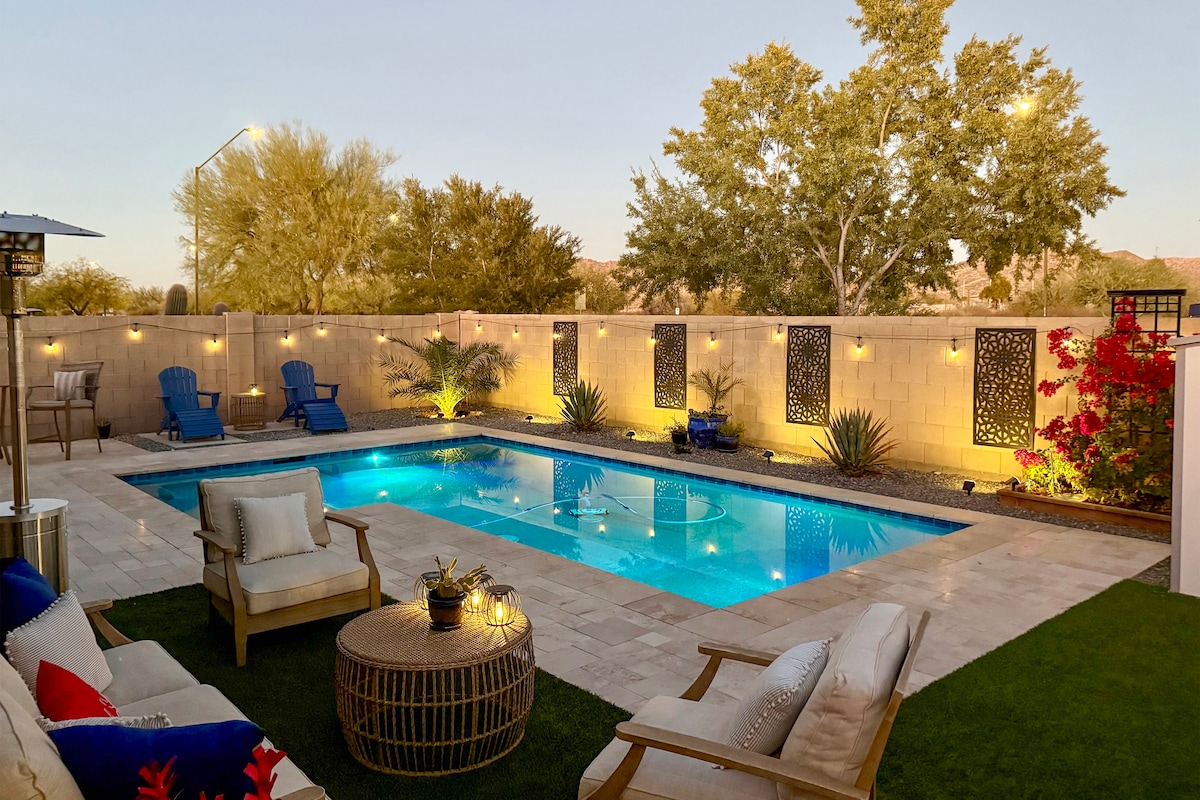 The outdoor area features a private pool surrounded by vibrant landscaping and illuminated by ambient lighting. Comfortably arranged seating, including lounge chairs and a cozy couch, offers a welcoming space for relaxation. A small table adds functionality to the serene patio environment.