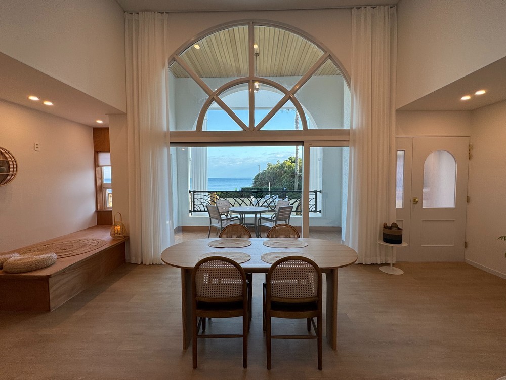 A spacious dining area is observed, featuring a round wooden table surrounded by rattan chairs. Large windows provide a view of the ocean, allowing natural light to fill the space. Soft curtains frame the windows, enhancing the open and airy feel of the room.