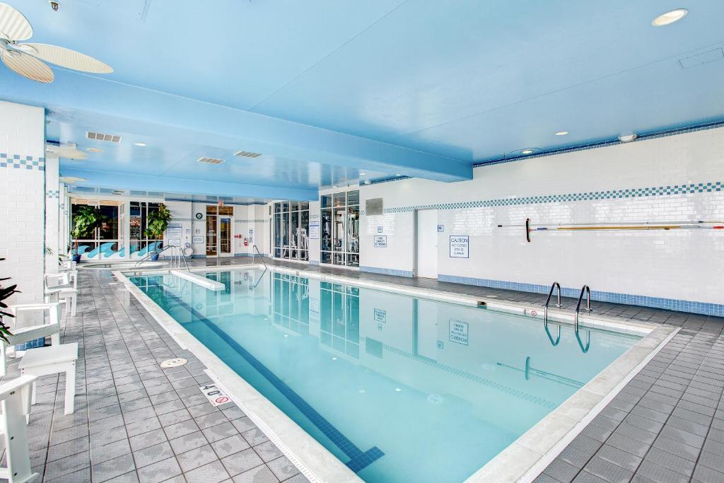 An indoor pool area features clear blue water, surrounded by tile flooring and brightly colored walls. Sunlight filters through large glass doors, illuminating the space. White lounge chairs line one side of the pool, providing a place for relaxation.