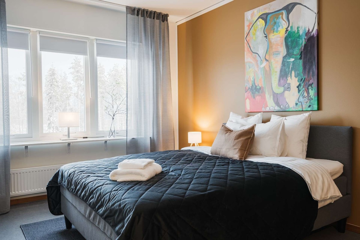 A serene bedroom features a spacious bed adorned with a black quilt and decorative pillows. The walls showcase a warm hue, complemented by large windows draped with light grey curtains. A piece of colorful artwork hangs above, while soft lighting creates a calm ambiance.