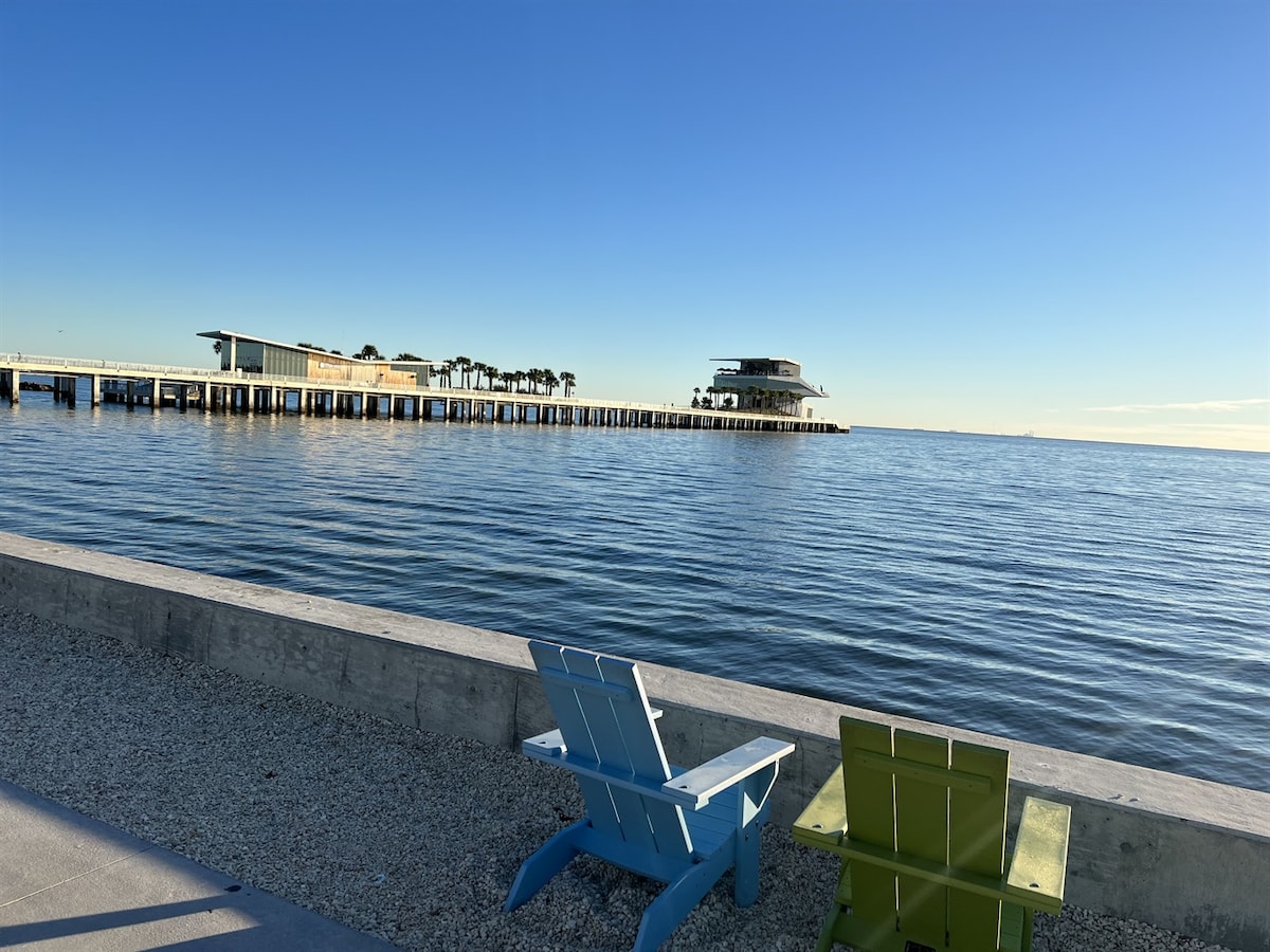 Two chairs in green and blue are positioned on a sandy area overlooking calm waters. The image features a distant pier with a modern structure, framed by a clear blue sky, enhancing the serene coastal ambiance.