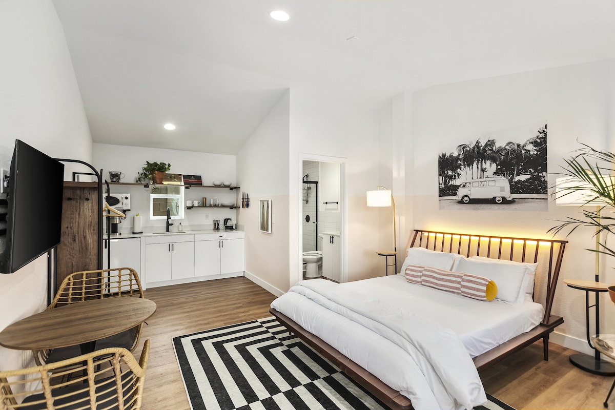 A modern studio space is presented, featuring a queen size bed with white linens and a decorative pillow. A kitchenette with white cabinetry is visible alongside a black and white striped rug. A wall-mounted television and two chairs are positioned for comfort, while a botanical element adds a touch of greenery.