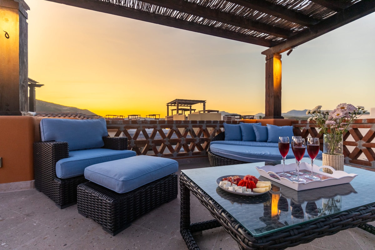 A rooftop terrace features cozy seating with plush blue cushions against a backdrop of a sunset sky. A glass table presents an assortment of desserts and glasses of red wine, complemented by a vase of flowers, creating a serene atmosphere for relaxation.