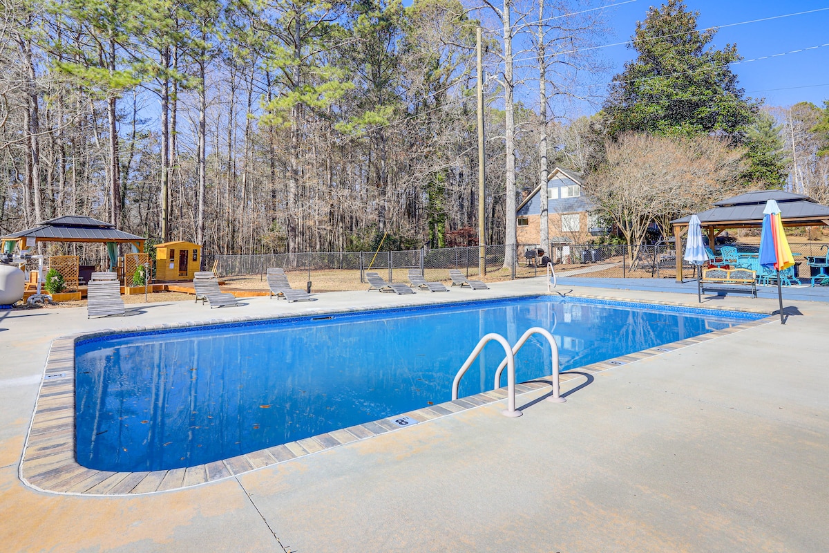 A large outdoor pool is surrounded by comfortable lounge chairs and shaded gazebos. Tall trees provide a natural backdrop, enhancing the serene atmosphere. The water's surface glistens in the sunlight, inviting refreshment on warm days.