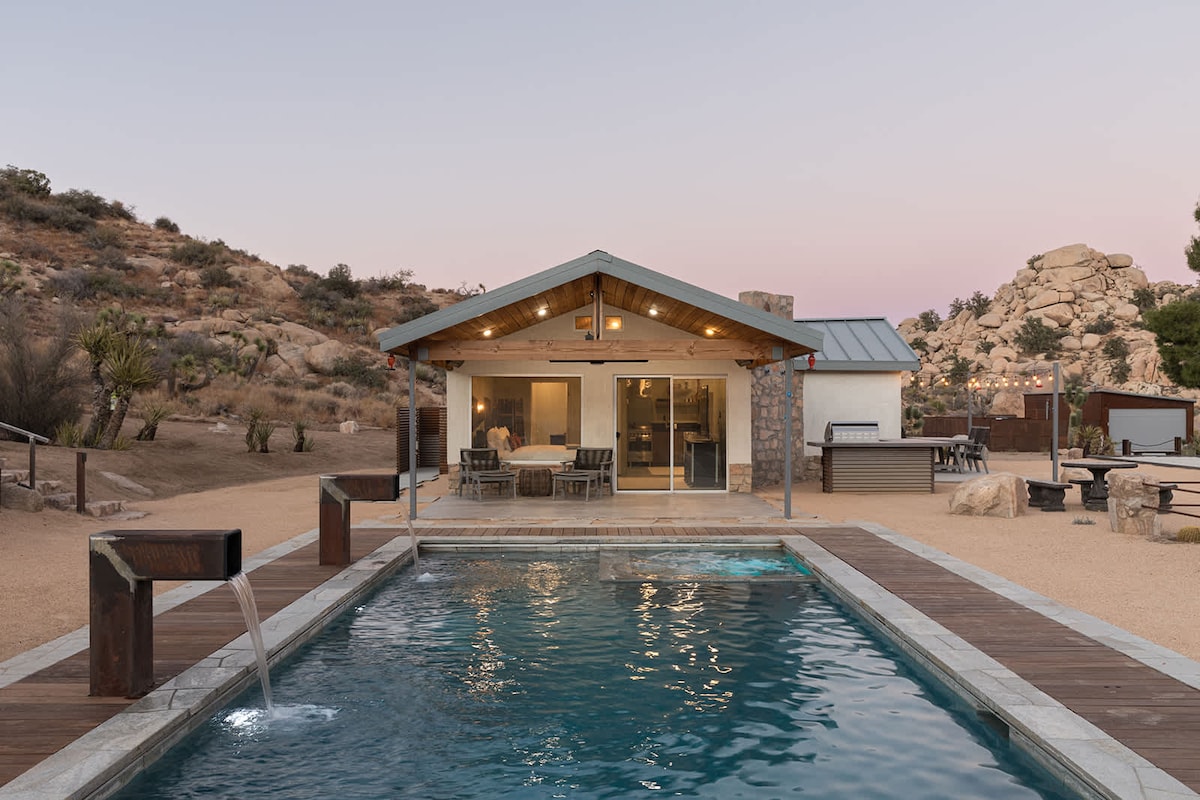 The outdoor area features an in-ground swimming pool with a fountain spillway. The modern structure has large glass doors leading to the interior. Surrounding the pool is a spacious deck, and natural desert landscape is visible in the background.