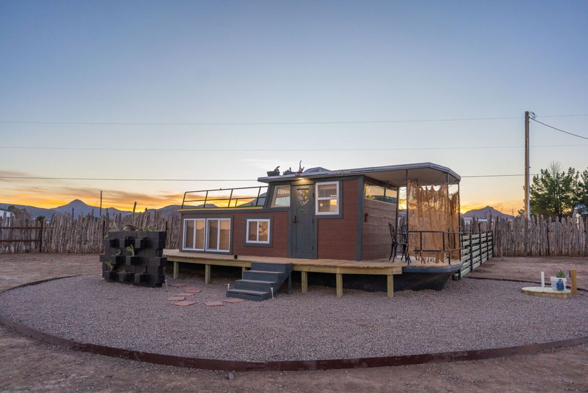 A renovated houseboat sits on a circular gravel pad, showcasing a spacious deck with seating and a scenic view of the sunset. The exterior features a mix of wood and durable materials, with large windows allowing for natural light. Surrounding the property is a rustic wooden fence.