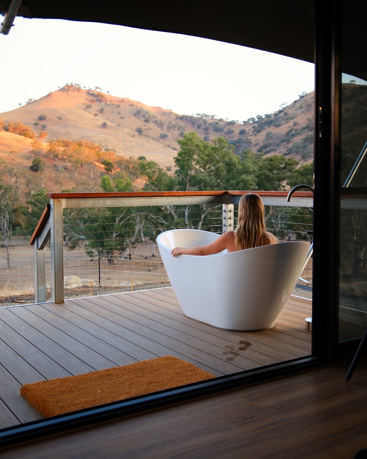 A freestanding white bathtub is positioned on a wooden deck, overlooking rolling hills in warm hues of orange and green. A person is seated in the tub, enjoying the serene landscape. The image is framed by an open sliding door, adding depth to the view.
