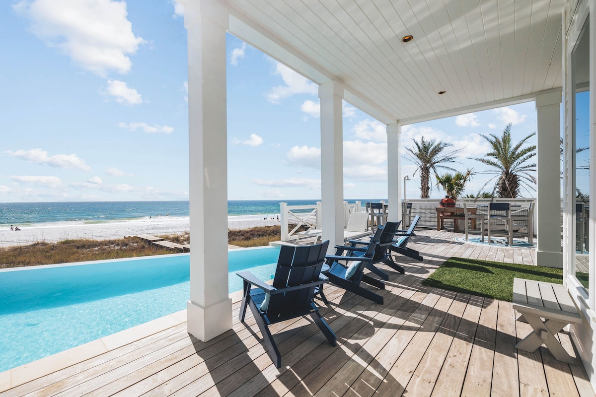 A spacious outdoor deck features comfortable blue chairs arranged to face the infinity pool and breathtaking Gulf views. Standing palm trees and sandy shoreline provide a picturesque backdrop, while the sunlit atmosphere invites relaxation and enjoyment by the water.