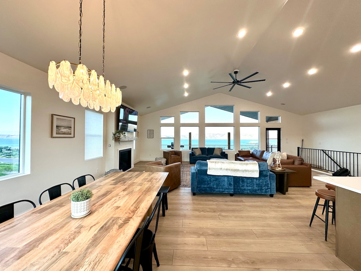 An open-concept living area features a large dining table and an inviting seating arrangement. Natural light floods through multiple windows, showcasing expansive views. A modern chandelier and ceiling fan add to the ambient feel. The space is designed for comfort and relaxation.