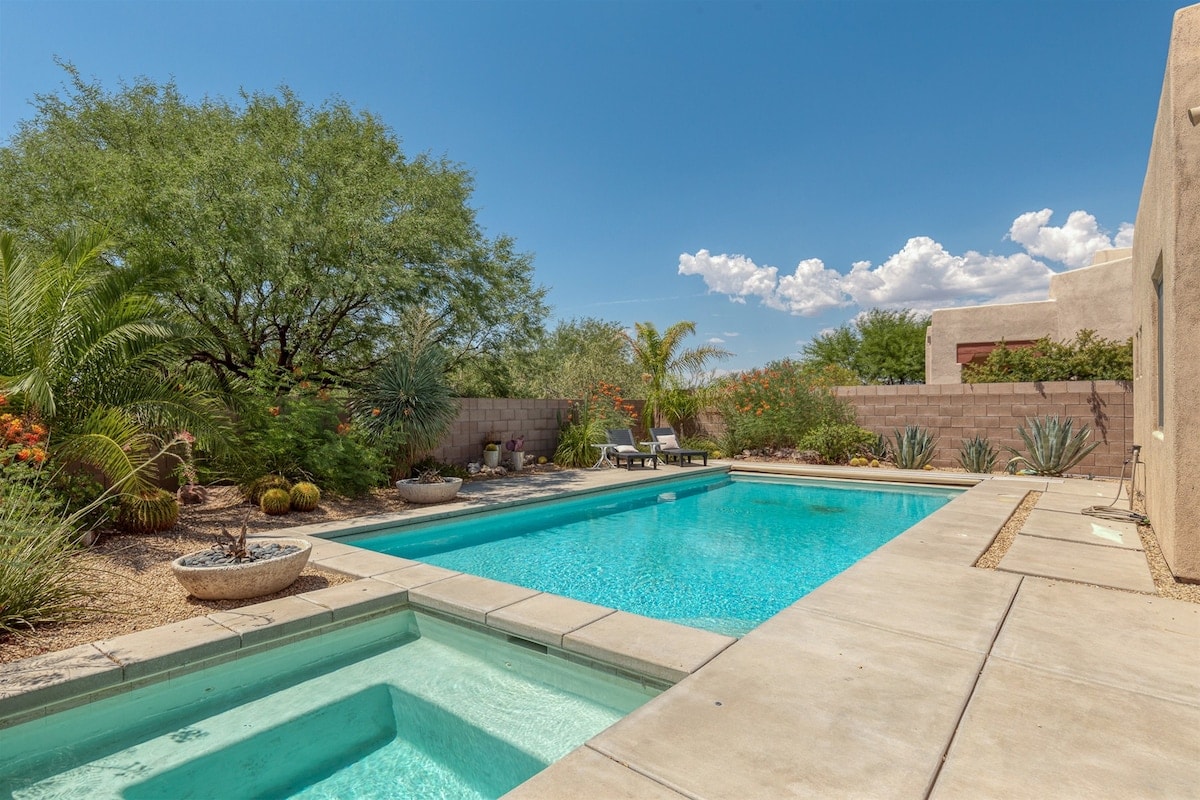 A serene backyard showcases a clear blue pool adjacent to a heated spa. Lush desert landscaping surrounds the area, featuring native plants and a soft stone pathway. Bright sky and fluffy clouds create a refreshing atmosphere, ideal for relaxation or outdoor activities.