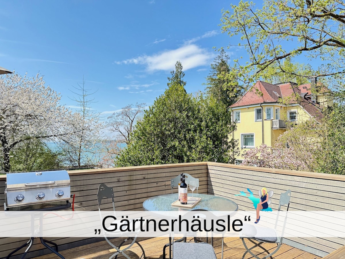 A spacious terrace is shown, featuring a dining table for four and a barbecue grill. Surrounding greenery adds a natural touch, while a glimpse of the lake can be seen in the background, complemented by the soft colors of blooming trees.