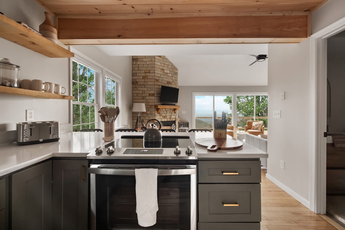 The kitchen features modern cabinetry with a stylish design, complemented by a sleek countertop. A stove is centered on the island, which is surrounded by utensils and cookware. Natural light fills the space through large windows, offering views of the outdoor scenery.