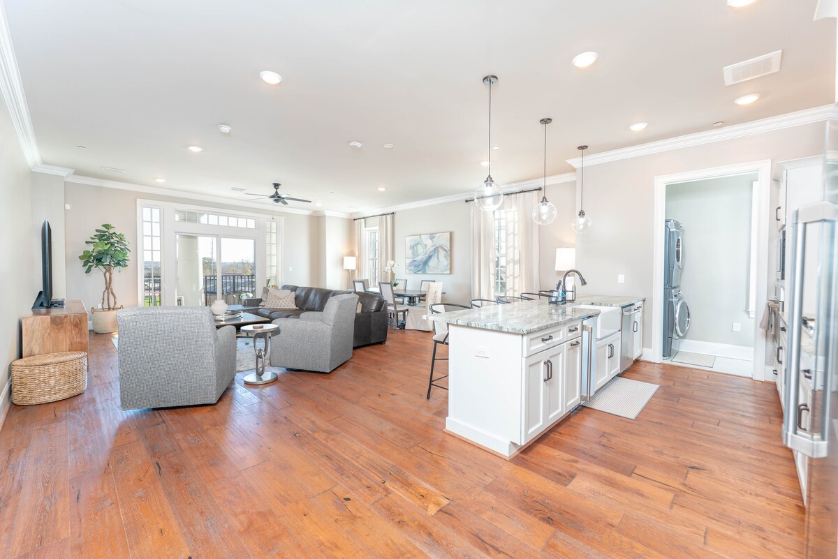 An airy living space features an open layout, with modern furnishings including two gray sofas and a coffee table. A well-appointed kitchen with a granite countertop complements the area. Natural light floods in through large windows, enhancing the inviting atmosphere.