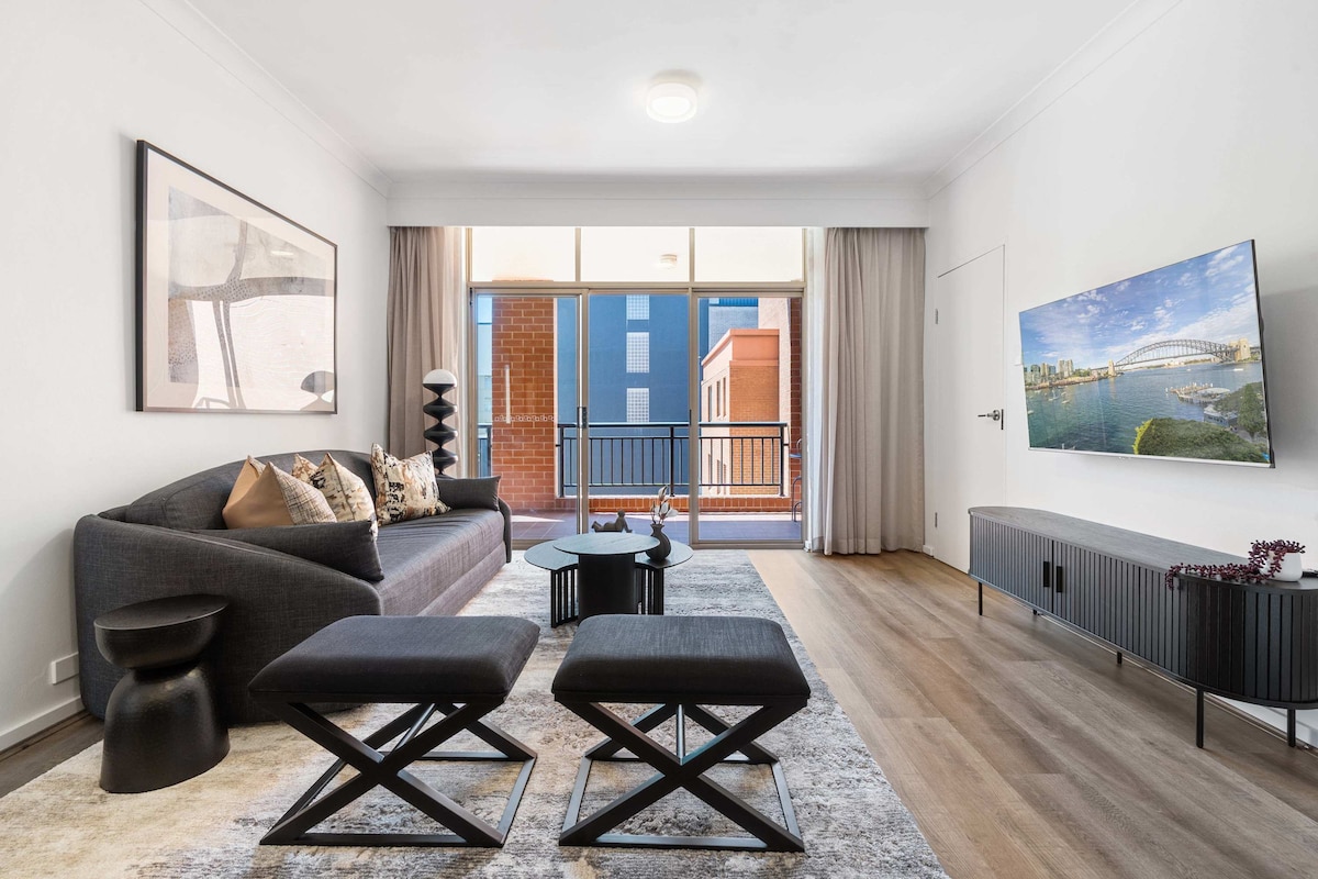 A modern living area features a spacious sectional sofa adorned with decorative cushions. A sleek TV mounted on the wall complements a low-profile media console. Natural light filters through large glass doors, which lead to a private balcony, enhancing the room's welcoming ambiance.