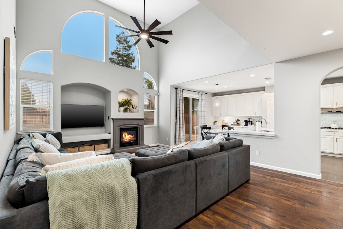 A spacious living area features a large sectional sofa arranged around a modern fireplace. Natural light enters through multiple arched windows, creating an airy atmosphere. The open layout connects to a kitchen visible in the background, enhancing the inviting space for relaxation and gatherings.