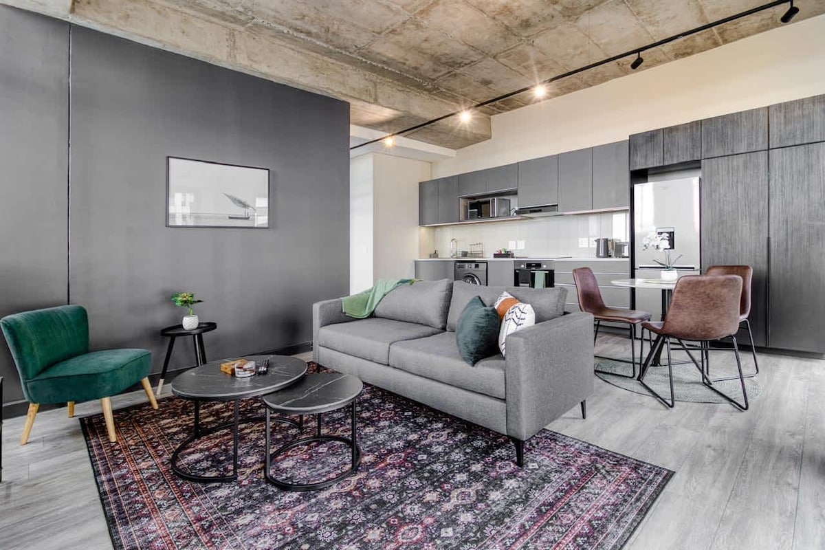 A modern living area features a gray sofa adorned with accent pillows, complemented by two stylish chairs and a patterned rug. A sleek dining table is visible with four chairs, while the kitchen area showcases contemporary cabinetry and appliances, creating a functional space for relaxation and dining.