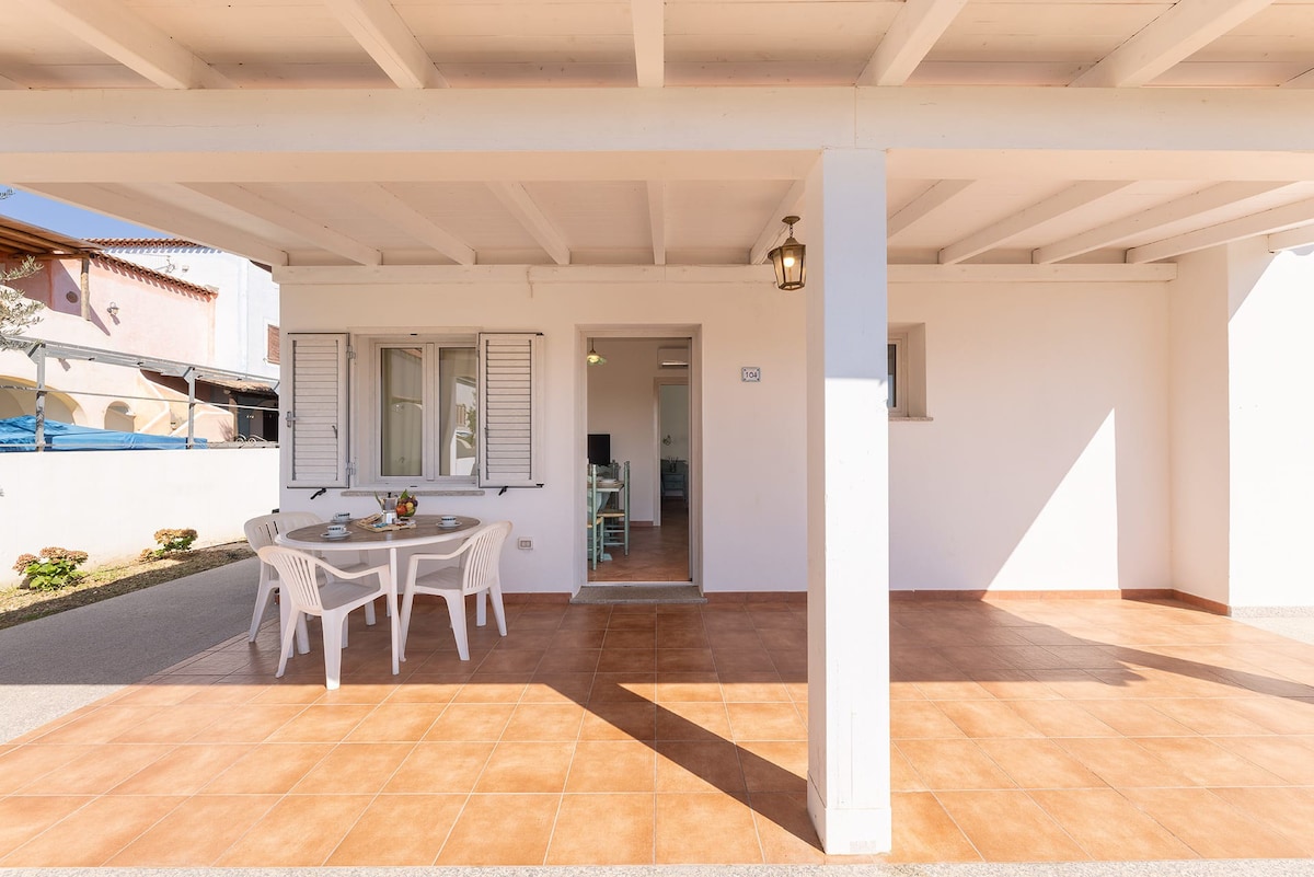 A covered outdoor area features a round table with four white chairs, set against tiled flooring. A doorway leads to the interior of the apartment, while natural light illuminates the space through nearby windows. The surrounding area includes well-maintained garden elements.