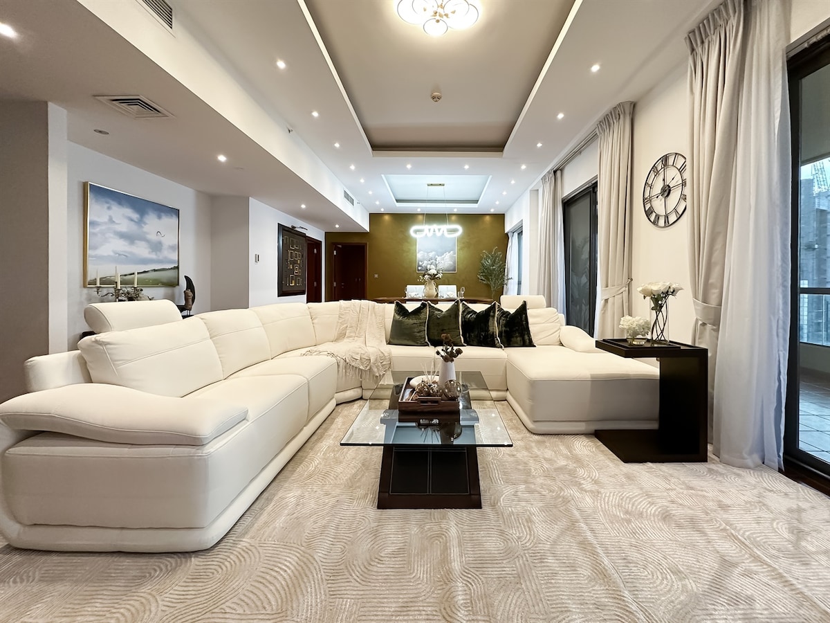 A spacious living area features a large white sectional sofa, elegantly arranged around a glass coffee table. Soft neutral tones decorate the space, complemented by natural light filtering through floor-to-ceiling windows. A decorative wall clock adds a touch of sophistication to the room.