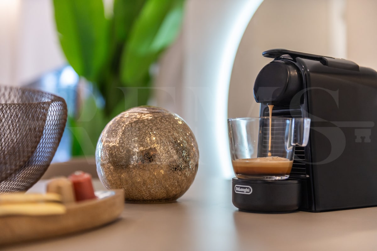 A sleek coffee machine is positioned on a surface, pouring a freshly brewed cup into a glass. A decorative sphere and a wicker basket add a touch of elegance, while lush green foliage is visible in the background, enhancing the modern aesthetic.
